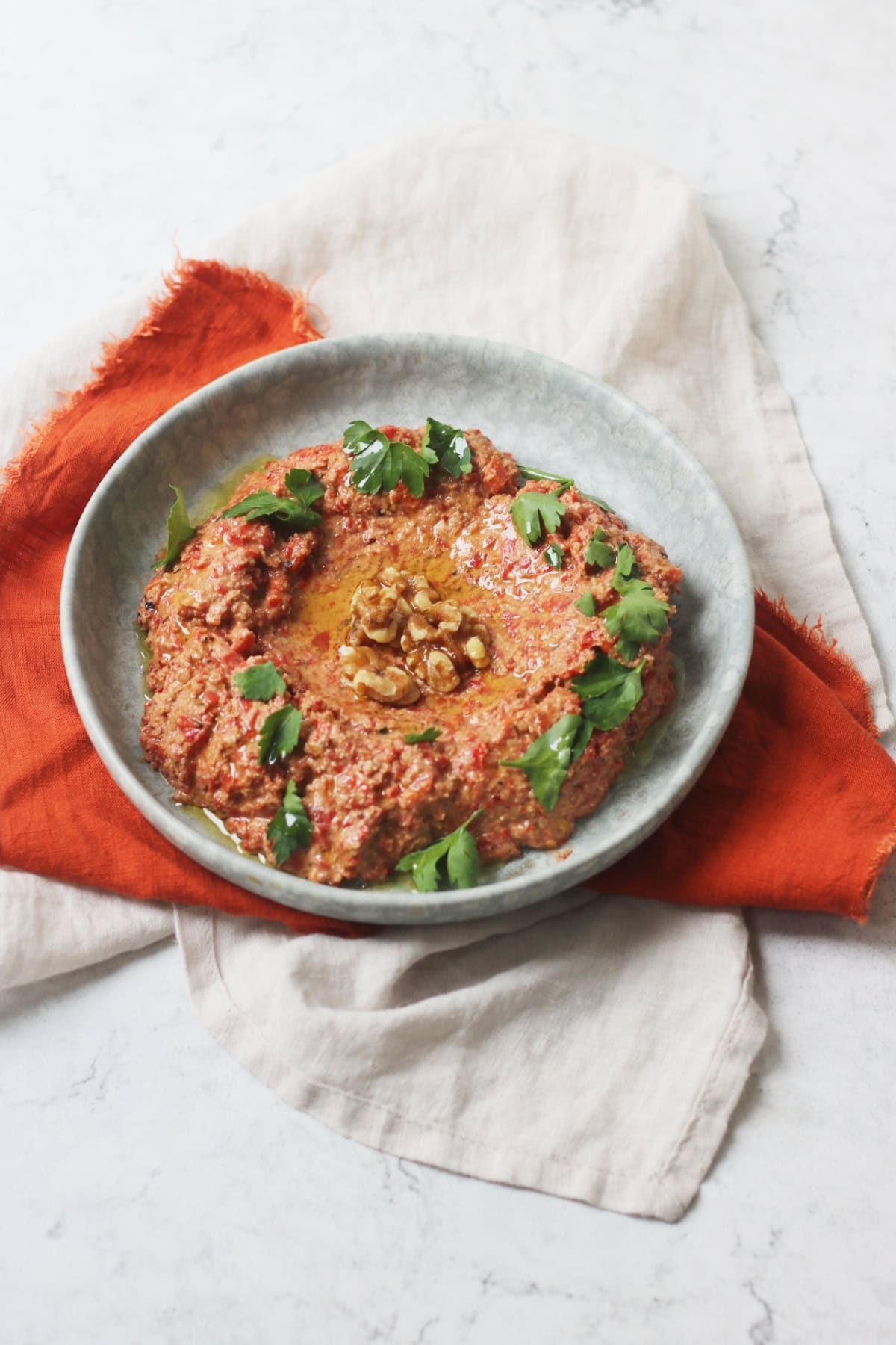 Muhammara, a roasted red pepper and walnut dip served with walnut halves and parsley on a pale blue plate with an orange napkin