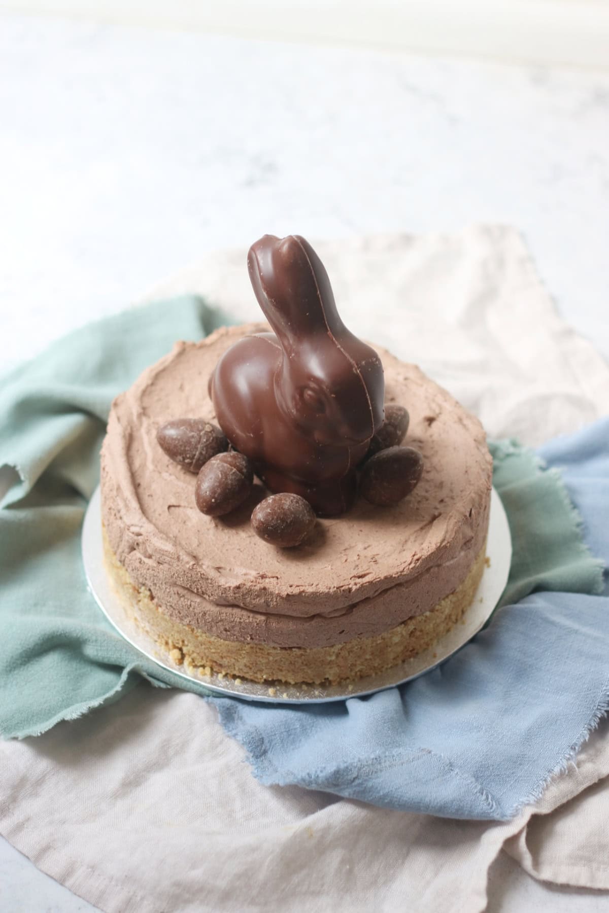 A Vegan Chocolate Cheesecake decorated with a chocolate bunny rabbit and small chocolate eggs sat on top of a silver cake board and blue and turquoise napkins