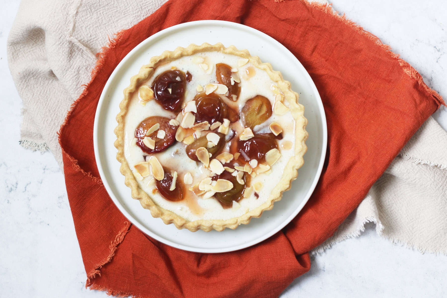 A vegan plum and custard tart topped with toasted flaked almonds on a white plate with a dark orange napkin.