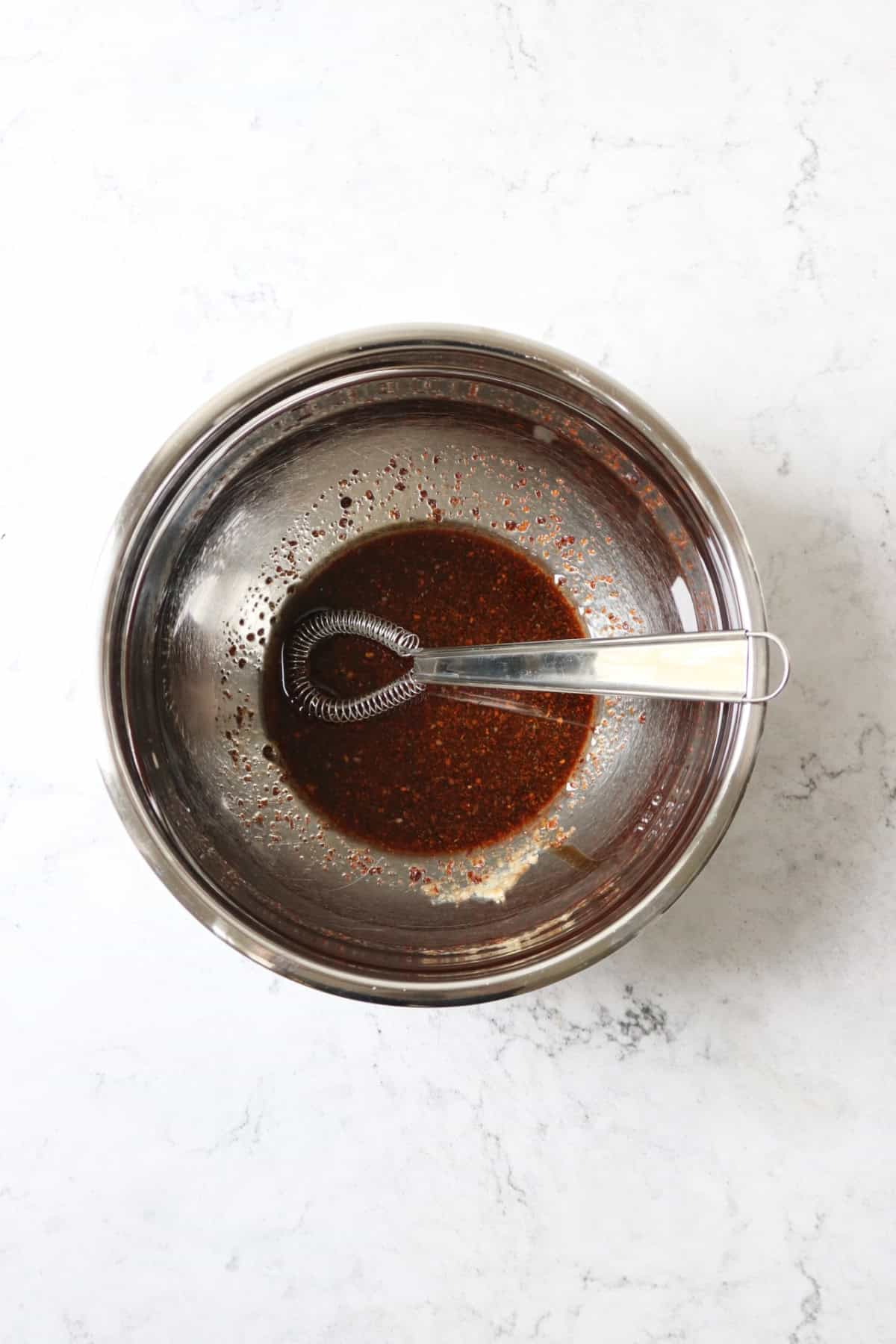 A metal mixing bowl containing a pomegranate molasses based marinade with a small sauce whisk