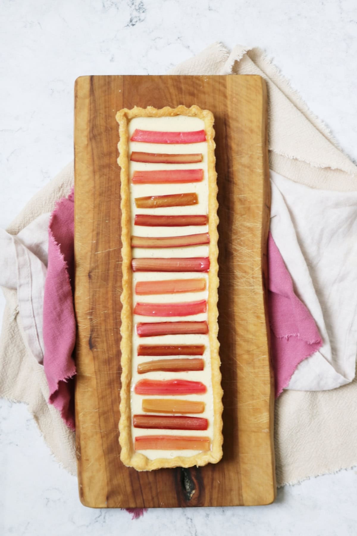 A rectangular Vegan Rhubarb and Custard Tart on a wooden serving board with pink and cream napkins underneath
