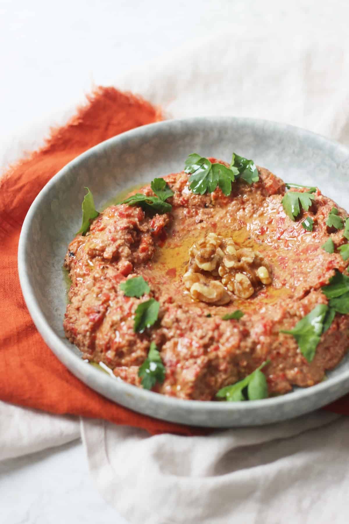 Muhammara, a roasted red pepper and walnut dip served with walnut halves and parsley on a pale blue plate with an orange napkin