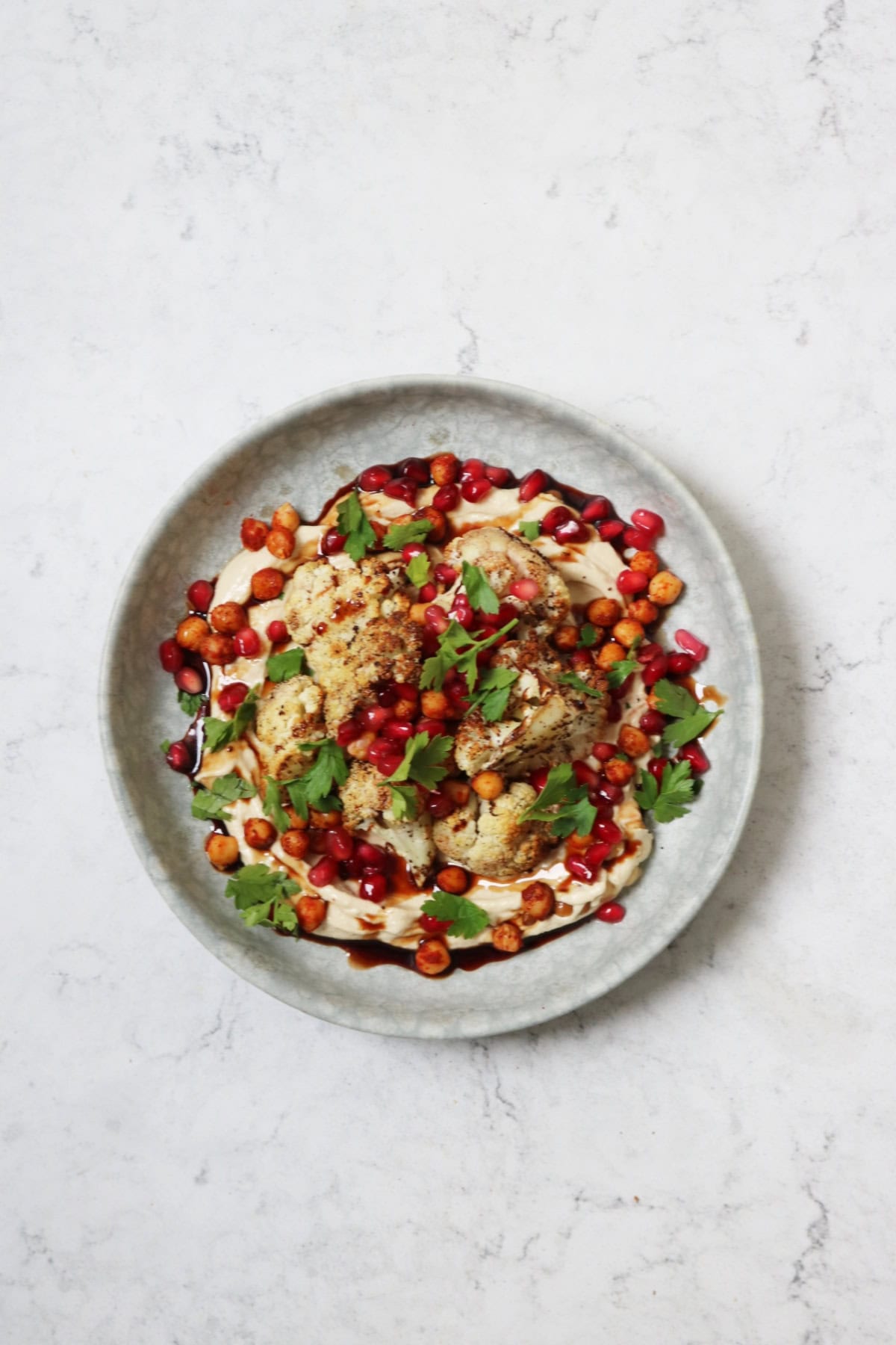 A plate of sumac roasted cauliflower on a bed of hummus with a drizzling of pomegranate molasses and roasted chickpeas, pomegranate seeds and parsley scattered over the top