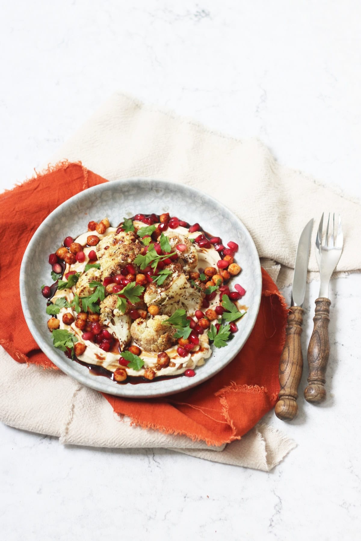 A plate of sumac roasted cauliflower on a bed of hummus with a drizzling of pomegranate molasses and roasted chickpeas, pomegranate seeds and parsley scattered over the top