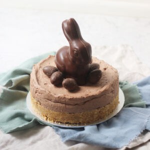 A Vegan Chocolate Cheesecake decorated with a chocolate bunny rabbit and small chocolate eggs sat on top of a silver cake board and blue and turquoise napkins