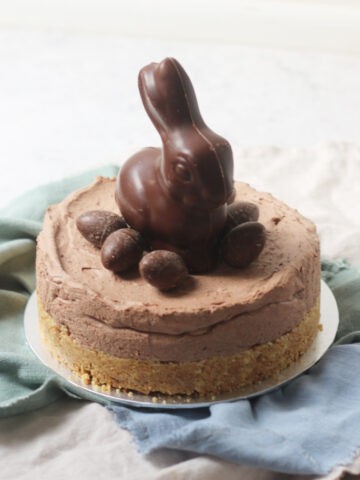 A Vegan Chocolate Cheesecake decorated with a chocolate bunny rabbit and small chocolate eggs sat on top of a silver cake board and blue and turquoise napkins