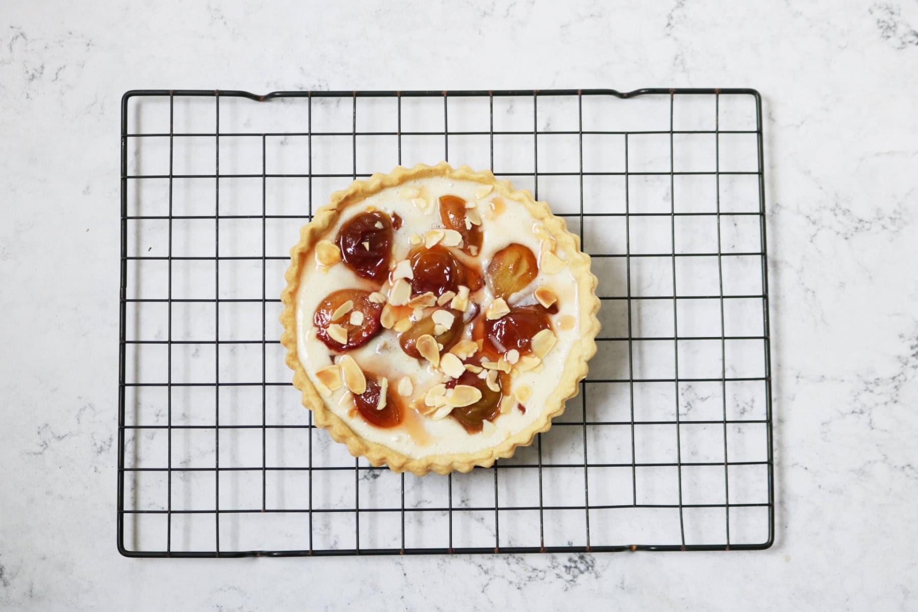 A vegan plum and custard tart topped with toasted flaked almonds sat on a black wire rack.