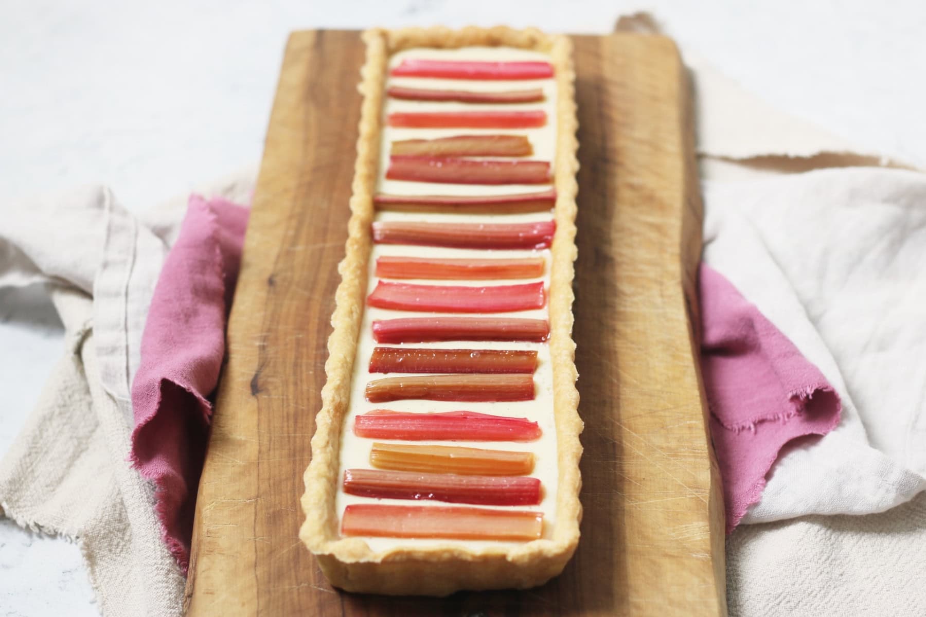 A rectangular Vegan Rhubarb and Custard Tart on a wooden serving board with pink and cream napkins underneath