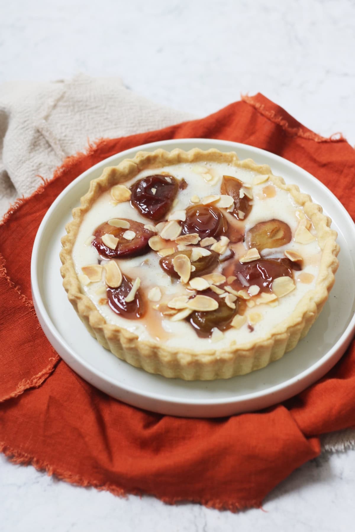 A vegan plum and custard tart topped with toasted flaked almonds on a white plate with a dark orange napkin.