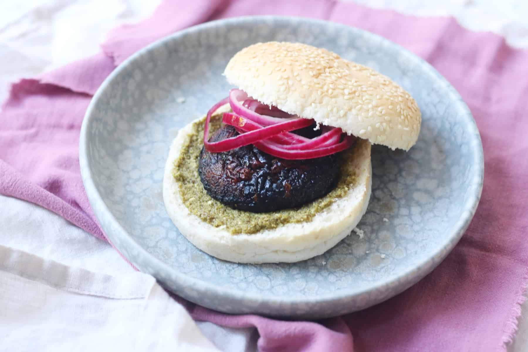 A BBQ Portobello Mushroom Burger in a bun with pesto and pickled red onion rings