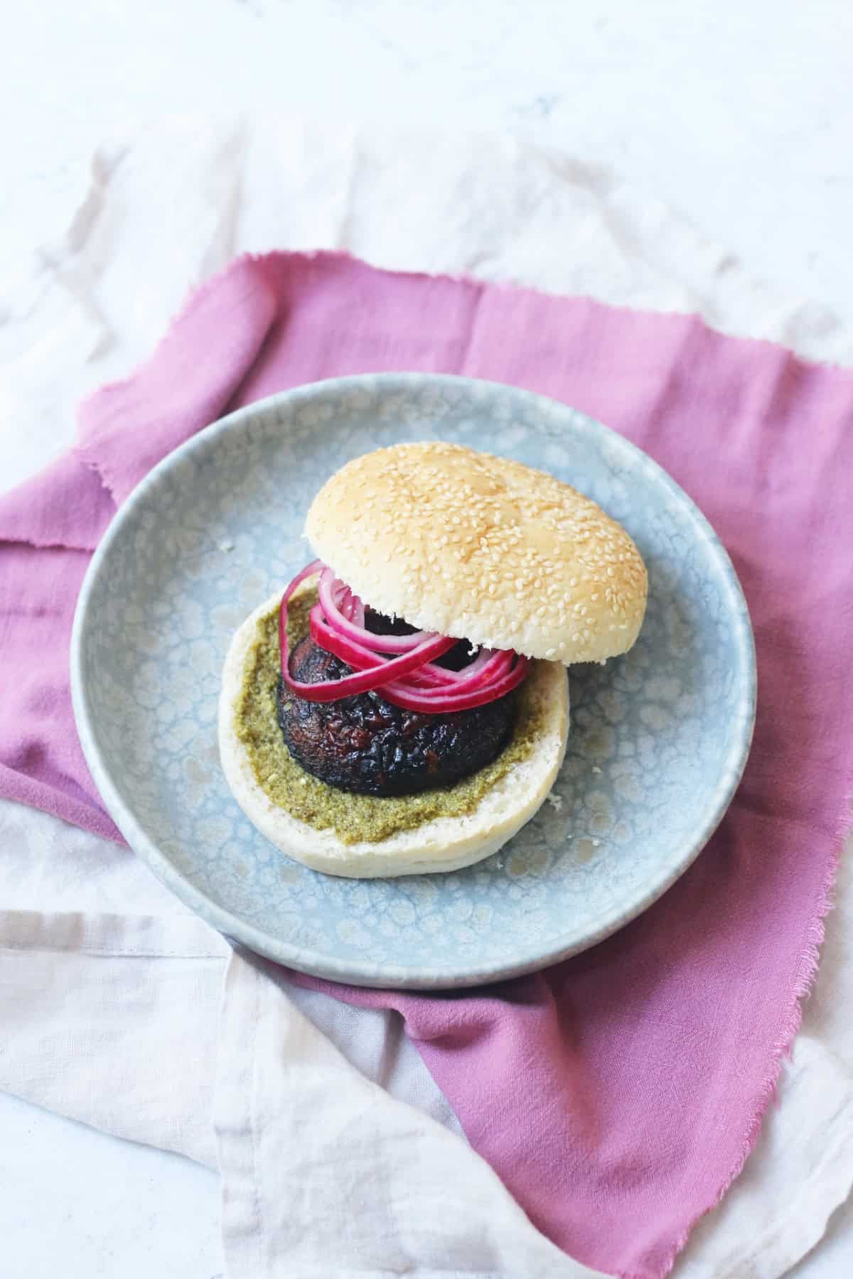 A BBQ Portobello Mushroom Burger in a bun with pesto and pickled red onion rings