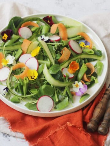 A vibrant Spring Garden Salad topped with edible flowers served on a white plate with an orange napkin and wooden handled knife and fork