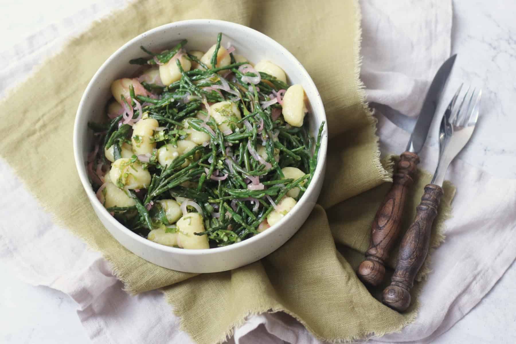 A bowl of Vegan Samphire Gnocchi served in a white bowl with a green napkin and wooden handled knife and fork
