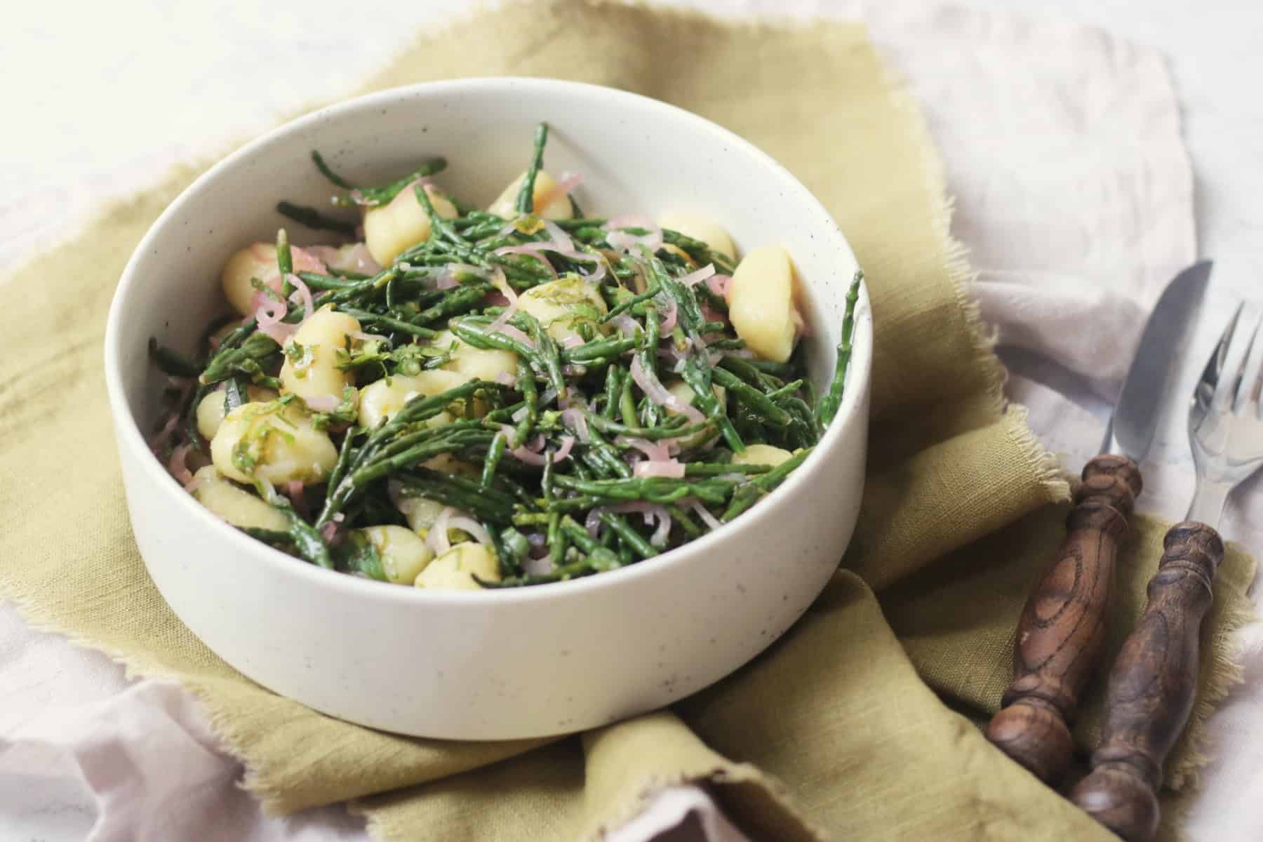 A bowl of Vegan Samphire Gnocchi served in a white bowl with a green napkin and wooden handled knife and fork