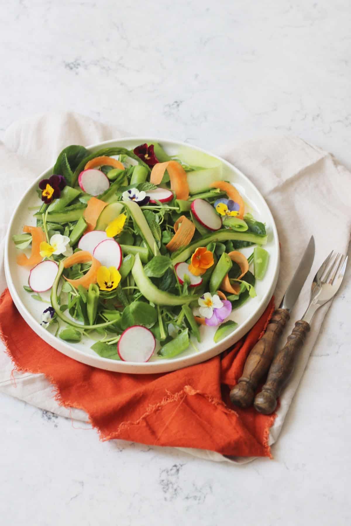 A vibrant Spring Garden Salad topped with edible flowers served on a white plate with an orange napkin and wooden handled knife and fork