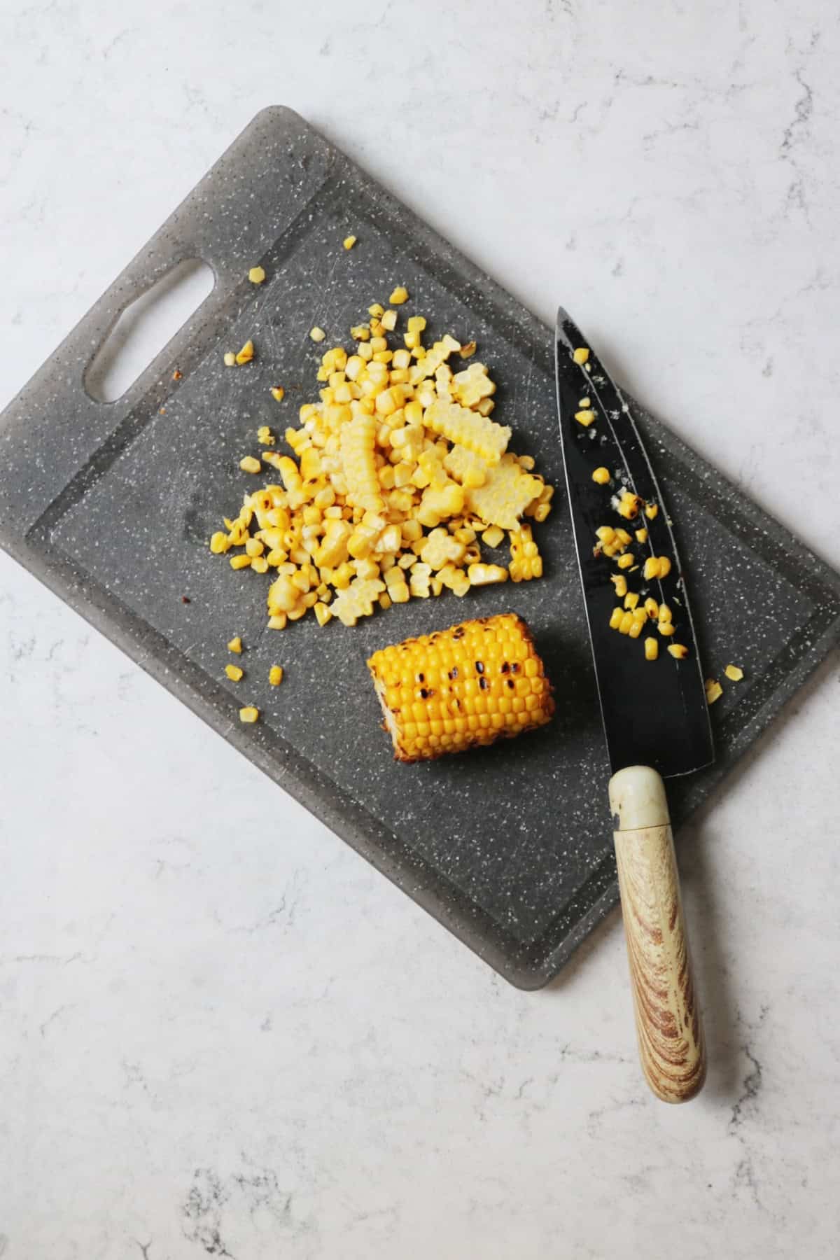 Kernels being cut from a corn on a cob with a sharp knife onto a grey chopping board