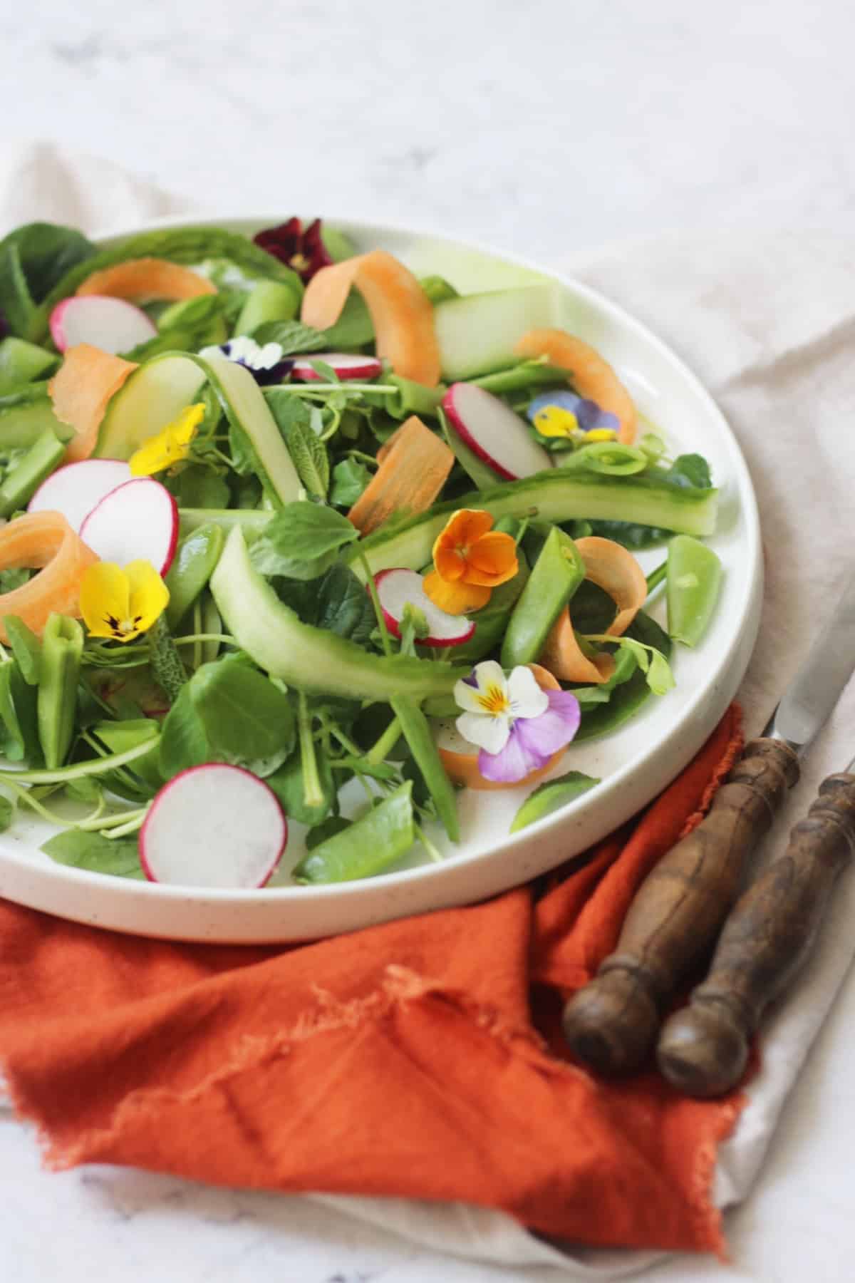 A vibrant Spring Garden Salad topped with edible flowers served on a white plate with an orange napkin and wooden handled knife and fork