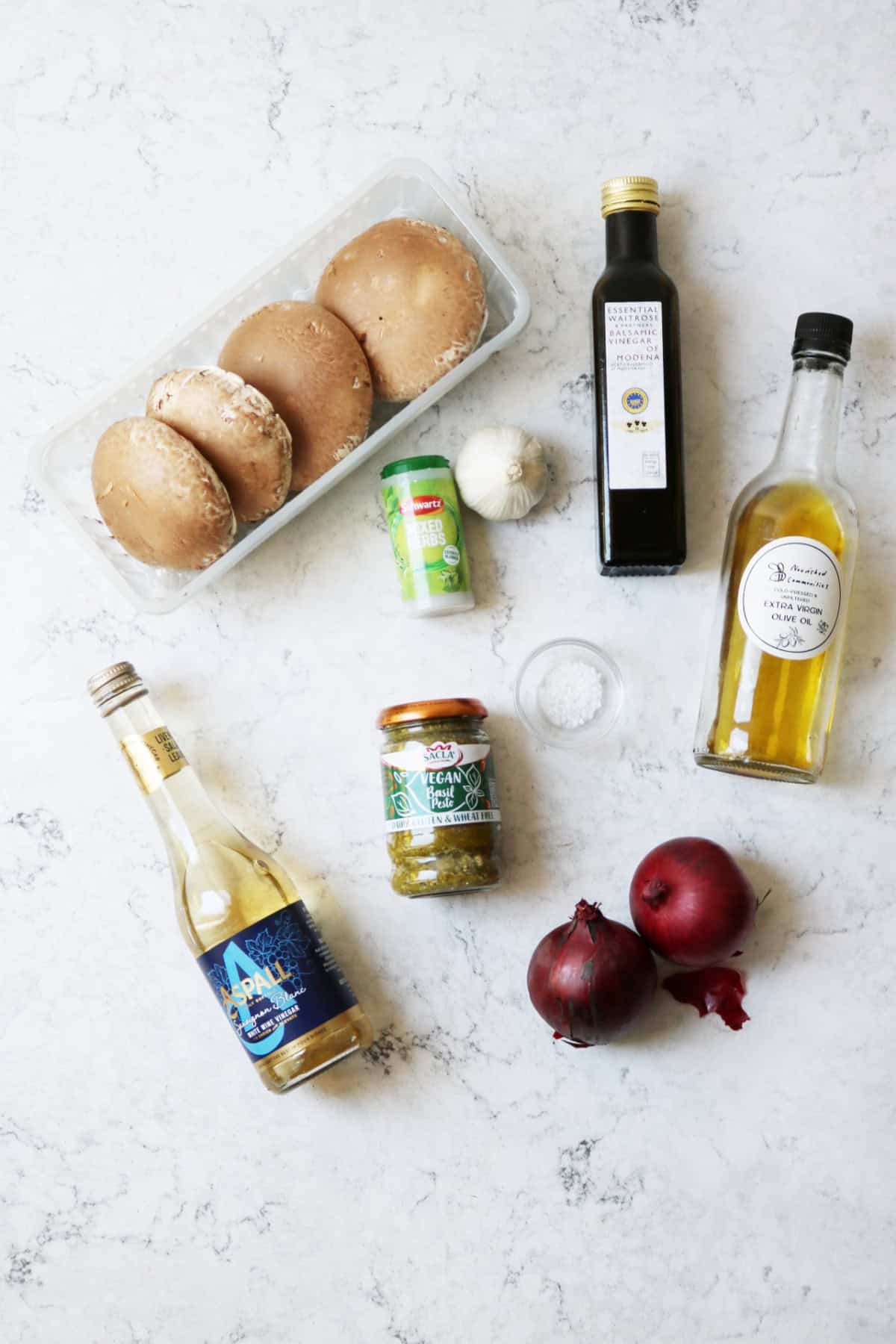 Portobello mushrooms, balsamic vinegar, mixed herbs, garlic, olive oil, white wine vinegar, pesto and red onions laid out on a white marble counter top