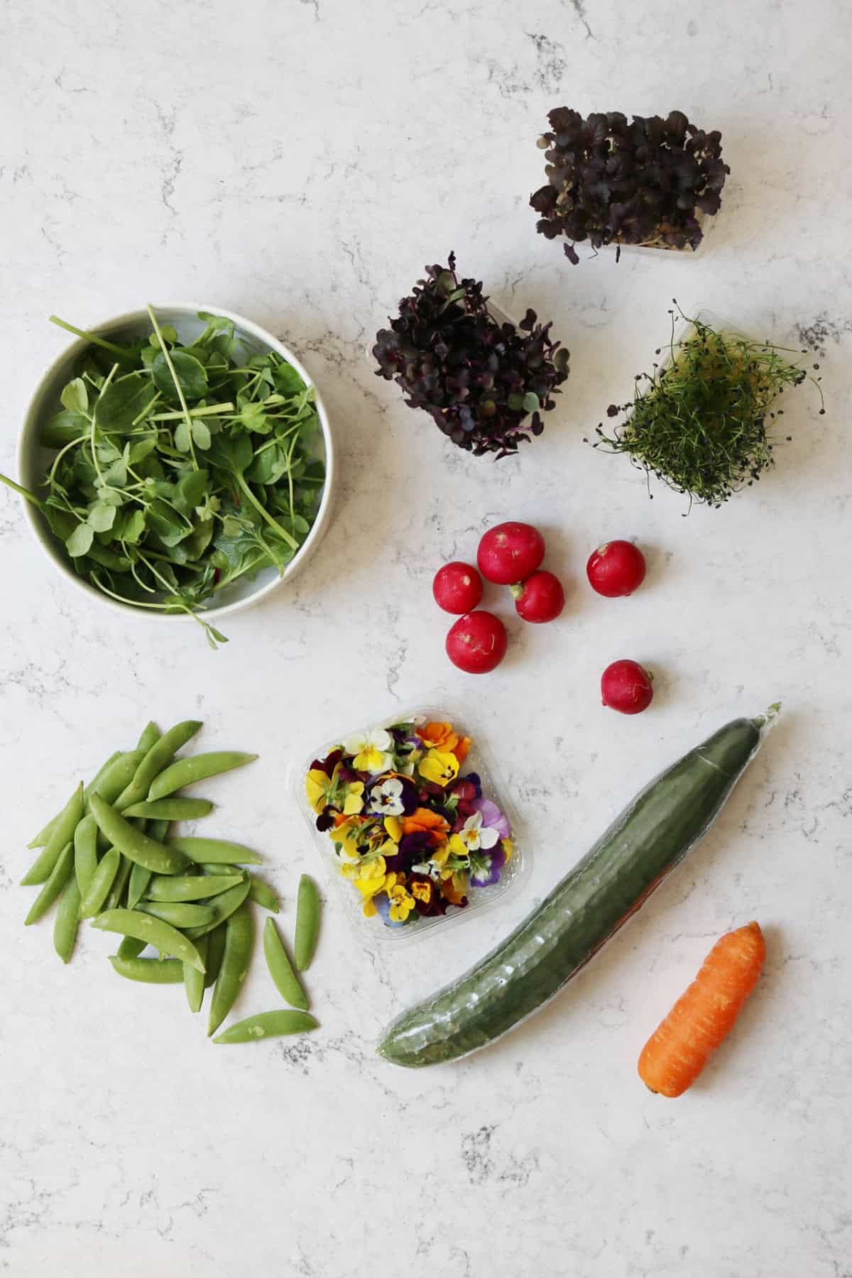 A bowl of pea shoots, micro herbs, sugar snap peas, edible flowers, radishes, a cucumber and a carrot laid out on a white marble counter top
