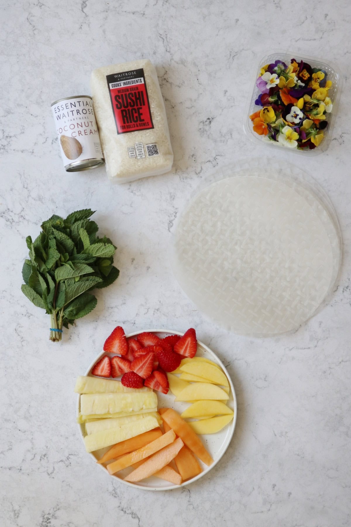 Coconut cream, sushi rice, edible flowers, mint leaves, rice paper wrappers and a selection of fruits laid out on a white marble counter top