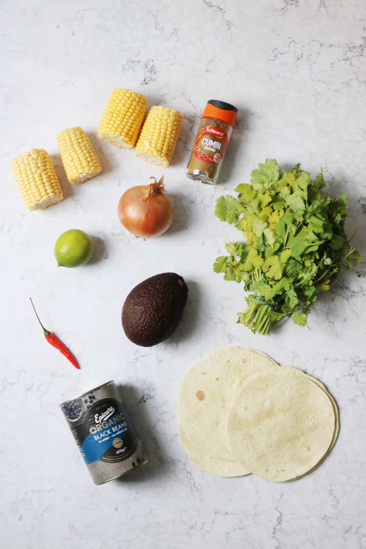 4 small corn on the cobs, ground cumin, a lime, an onion, a bunch of fresh coriander, a red chilli, an avocado, a tin of blacked beans and small flour tortillas laid out on a white marble countertop