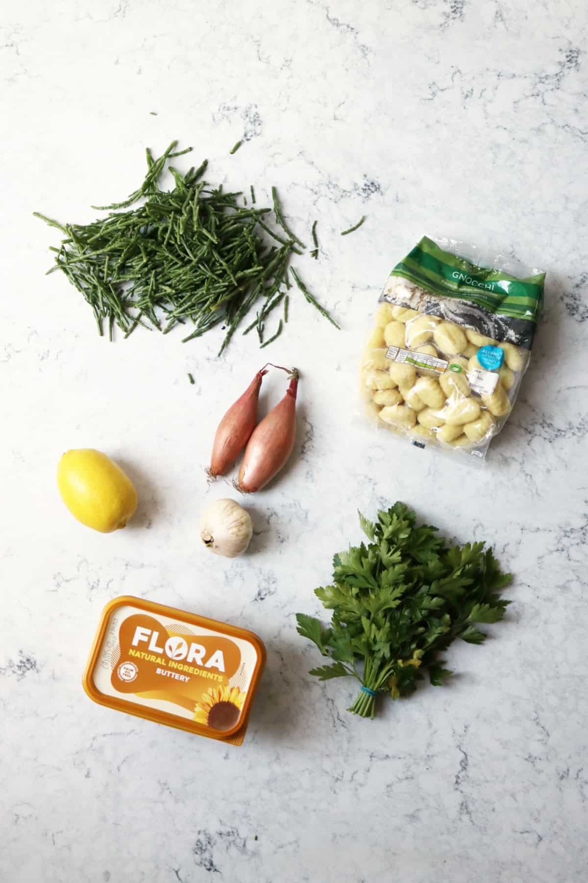 Samphire, a packet of gnocchi, a lemon, a bulb of garlic, a tub of vegan butter and a bunch of fresh parsley laid out on a white marble countertop