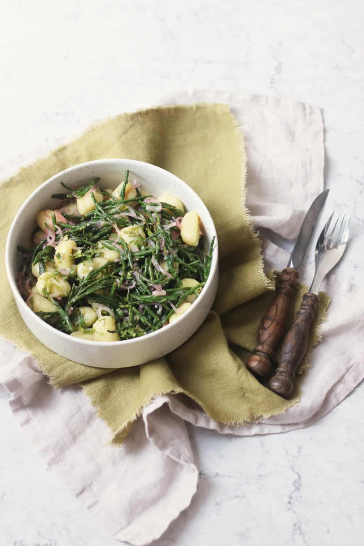 A bowl of Vegan Samphire Gnocchi served in a white bowl with a green napkin and wooden handled knife and fork