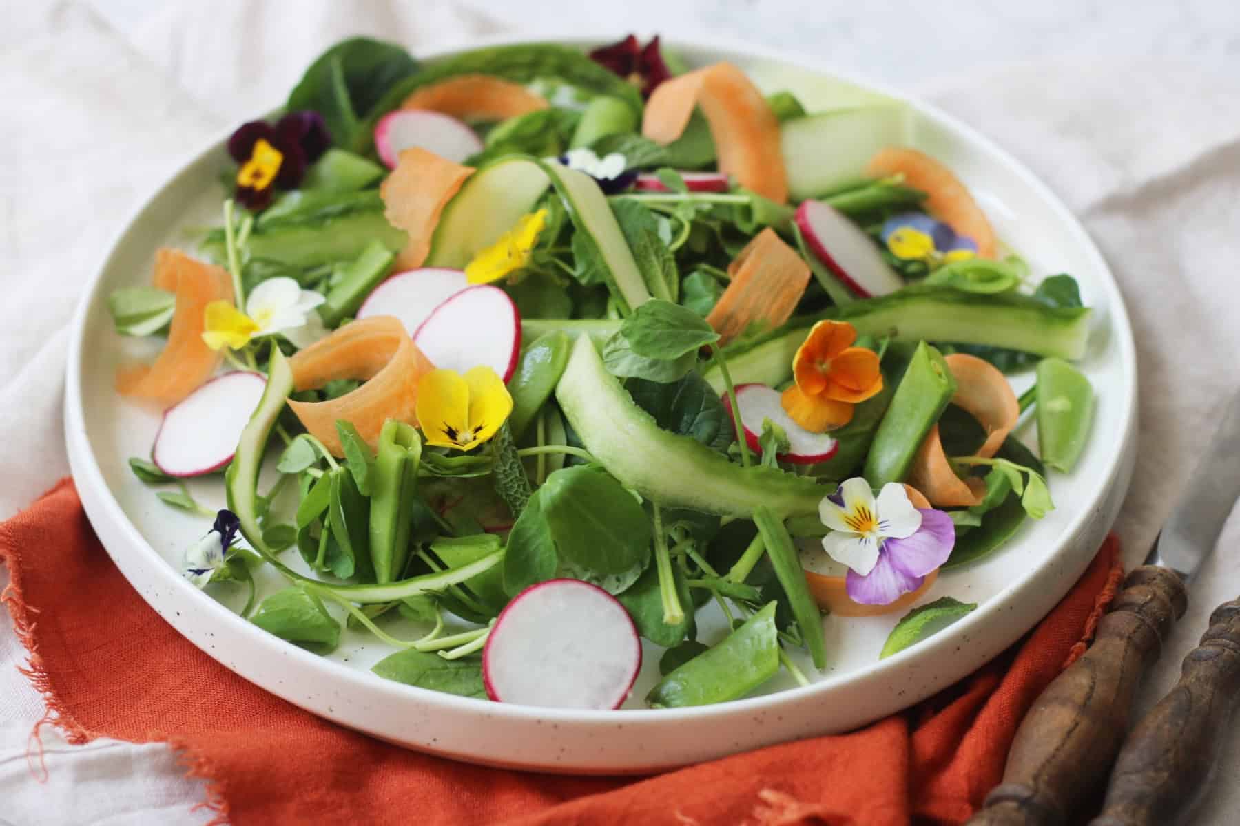 A vibrant Spring Garden Salad topped with edible flowers served on a white plate with an orange napkin and wooden handled knife and fork