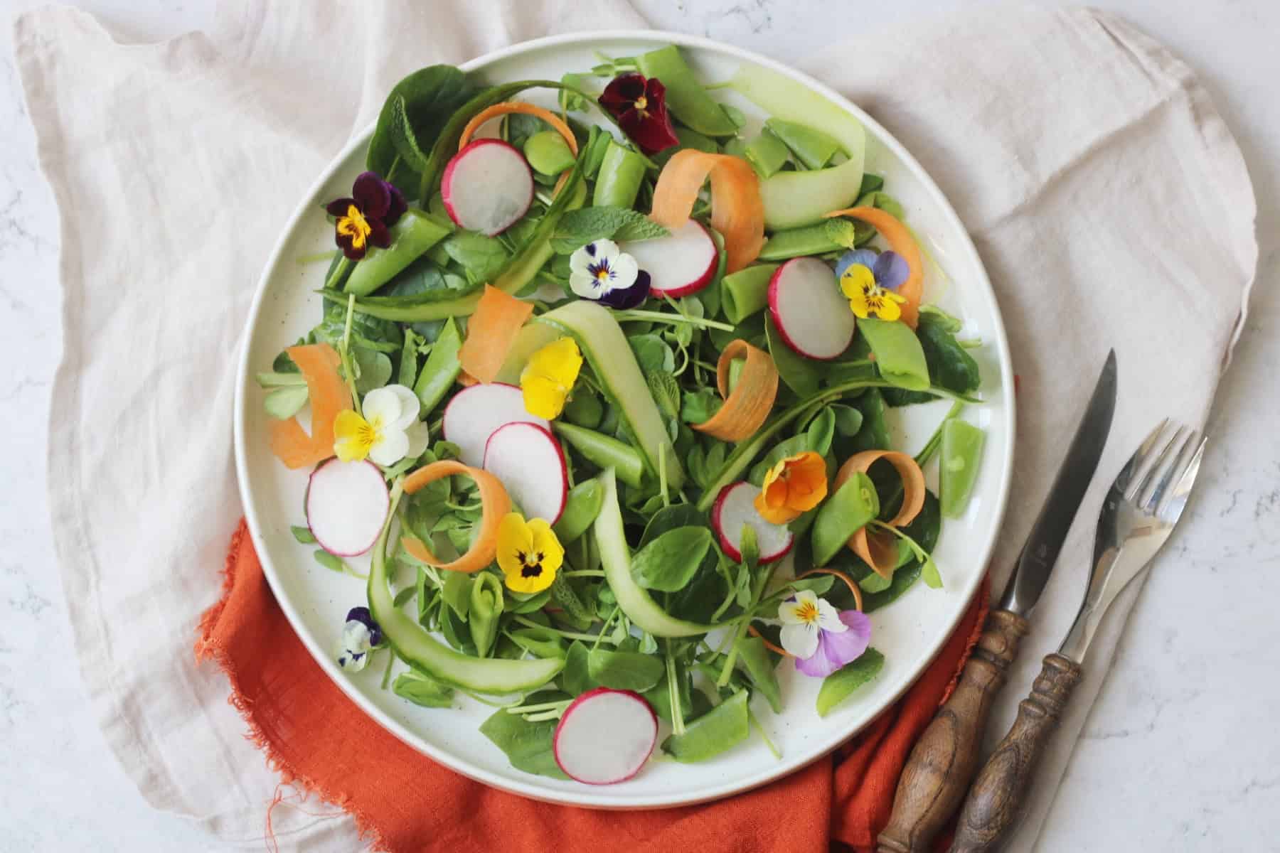A vibrant Spring Garden Salad topped with edible flowers served on a white plate with an orange napkin and wooden handled knife and fork