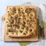 A Green Olive and Za'atar Focaccia on a wooden chopping board with a large bread knife