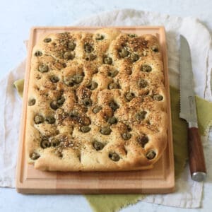 A Green Olive and Za'atar Focaccia on a wooden chopping board with a large bread knife