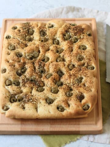 A Green Olive and Za'atar Focaccia on a wooden chopping board with a large bread knife