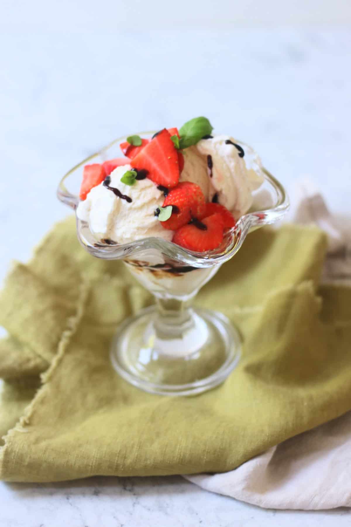 A Vegan Strawberry and Balsamic Ice Cream Sundae made up of layers of balsamic glaze, dairy free ice cream, macerated strawberries and basil leaves served in an ice cream sundae glass on top of a green napkin.