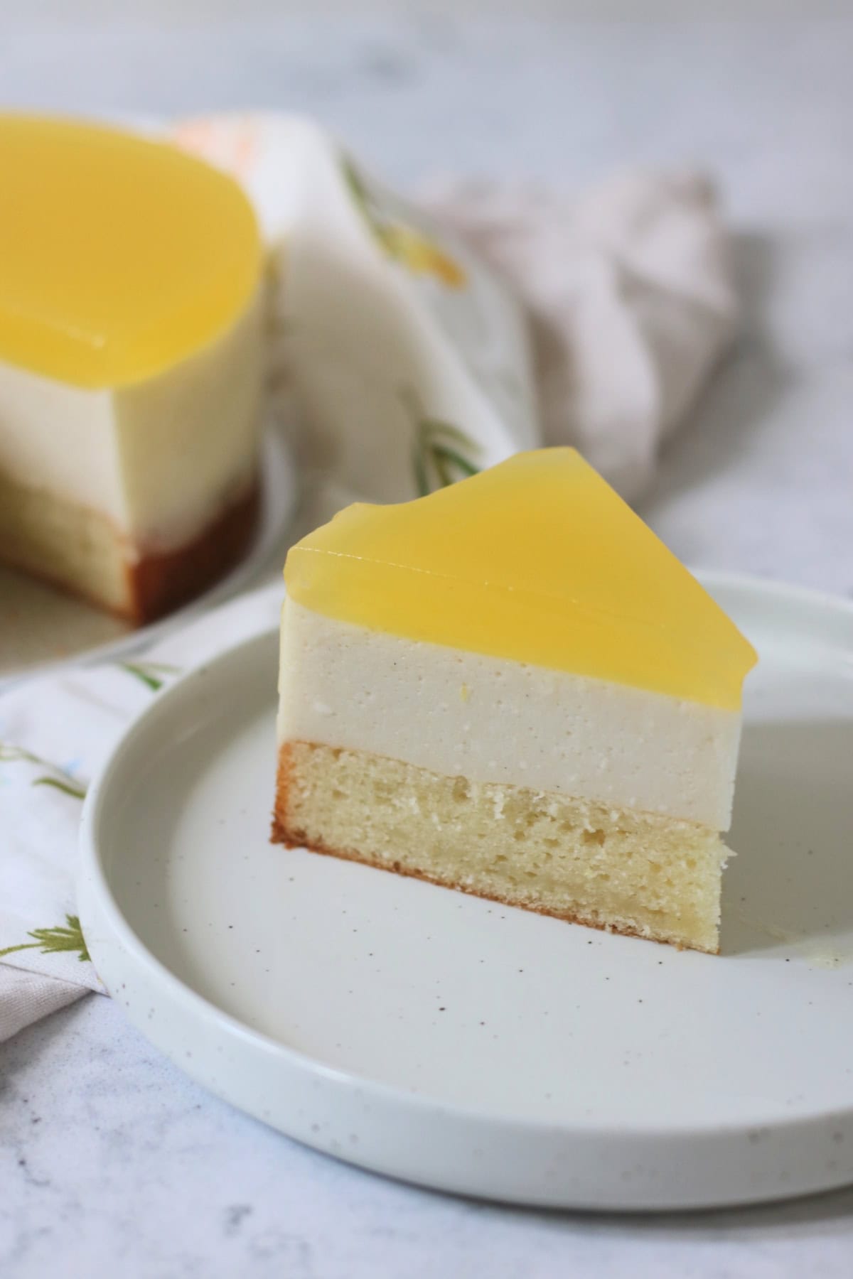 A large slice of Vegan White Chocolate and Lemon Mousse Cake showing layers of vanilla sponge, white chocolate and lemon mousse topped with lemon jelly.