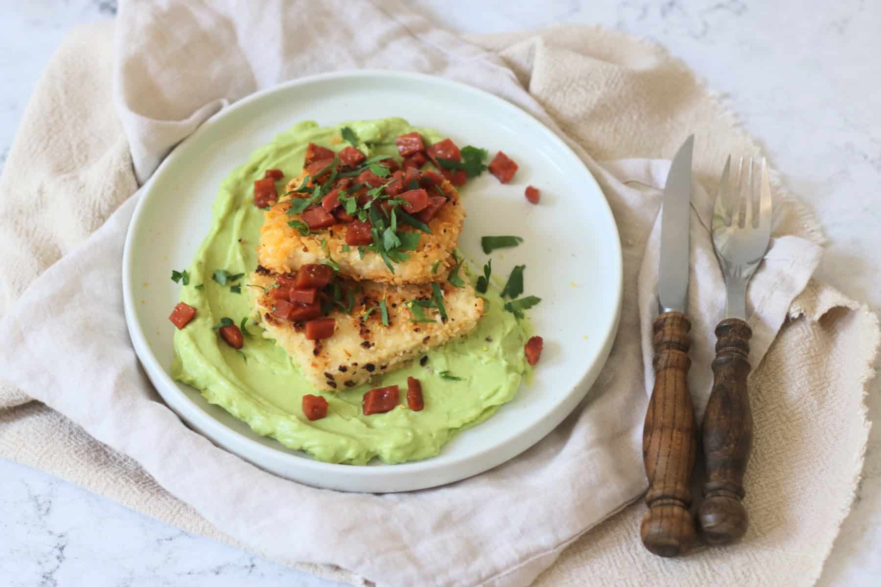 A plate of Crispy Lemon Pepper Tofu with Creamy Avocado Sauce and Bacon Crunch with fresh parsley and a wooden handled knife and fork.