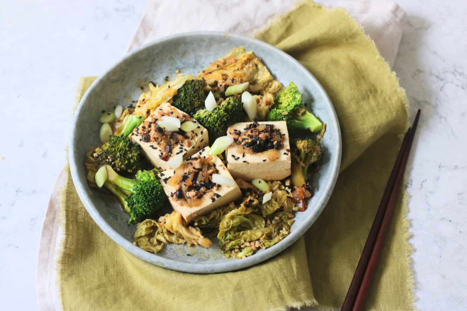3 pieces of Braised Black Bean Stuffed Tofu served with cabbage and broccoli on a plate with a green napkin and wooden chopsticks