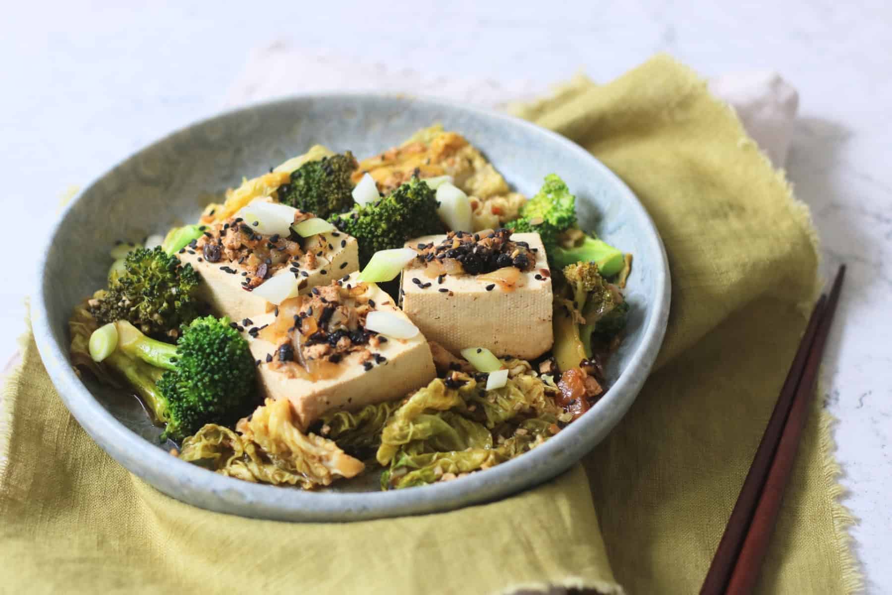 3 pieces of Braised Black Bean Stuffed Tofu served with cabbage and broccoli on a plate with a green napkin and wooden chopsticks