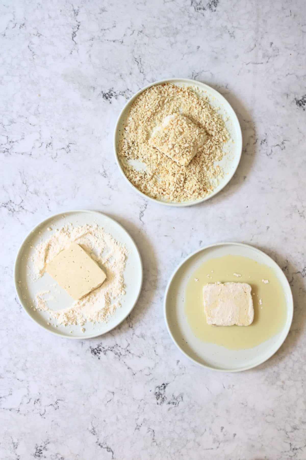 One slice of tofu being dipped in plant milk, one slice of tofu being dipped in flour and one slice of tofu being dipper in lemon-pepper breadcrumbs.