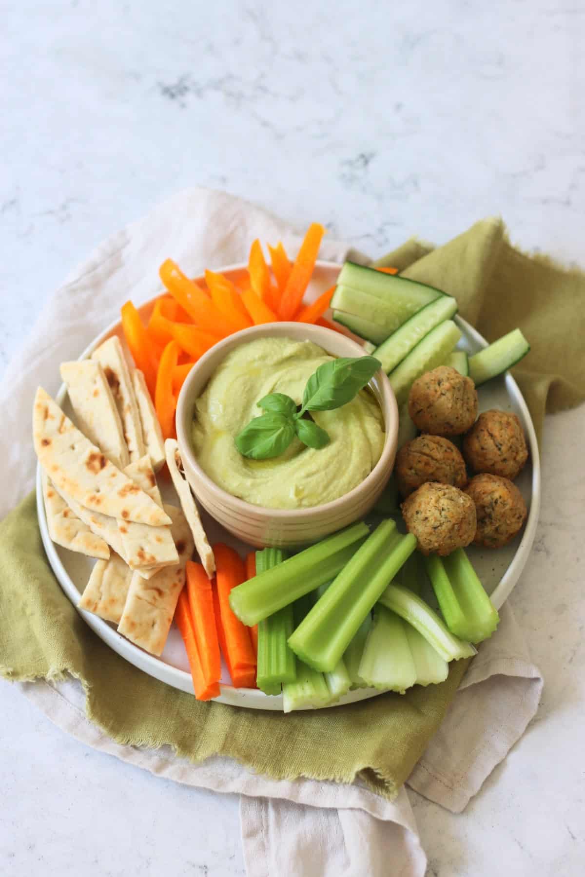 A crudites platter made up of pitta, carrots, celery, falafel, cucumber and orange pepper with a bowl of Roasted Garlic and Basil Hummus