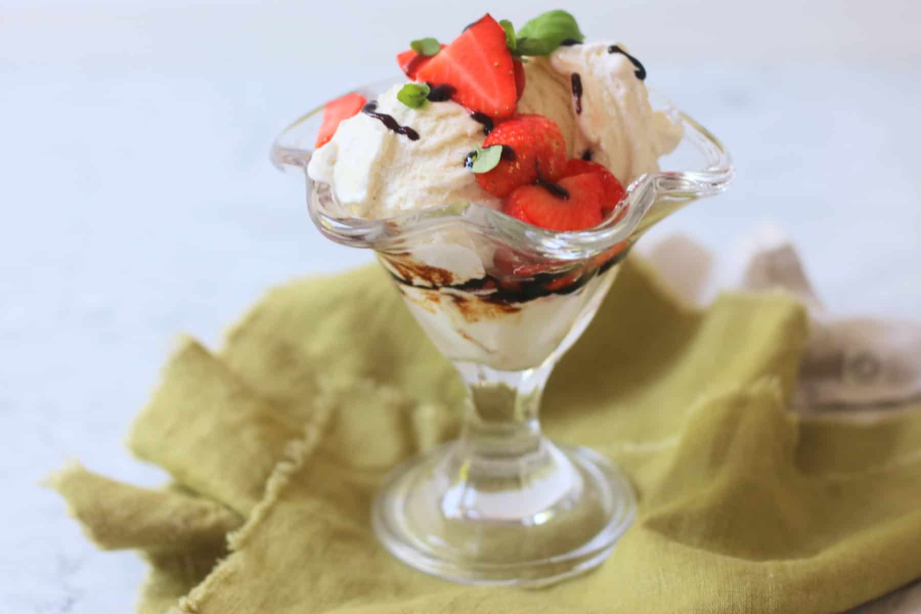 A Vegan Strawberry and Balsamic Ice Cream Sundae made up of layers of balsamic glaze, dairy free ice cream, macerated strawberries and basil leaves served in an ice cream sundae glass on top of a green napkin.