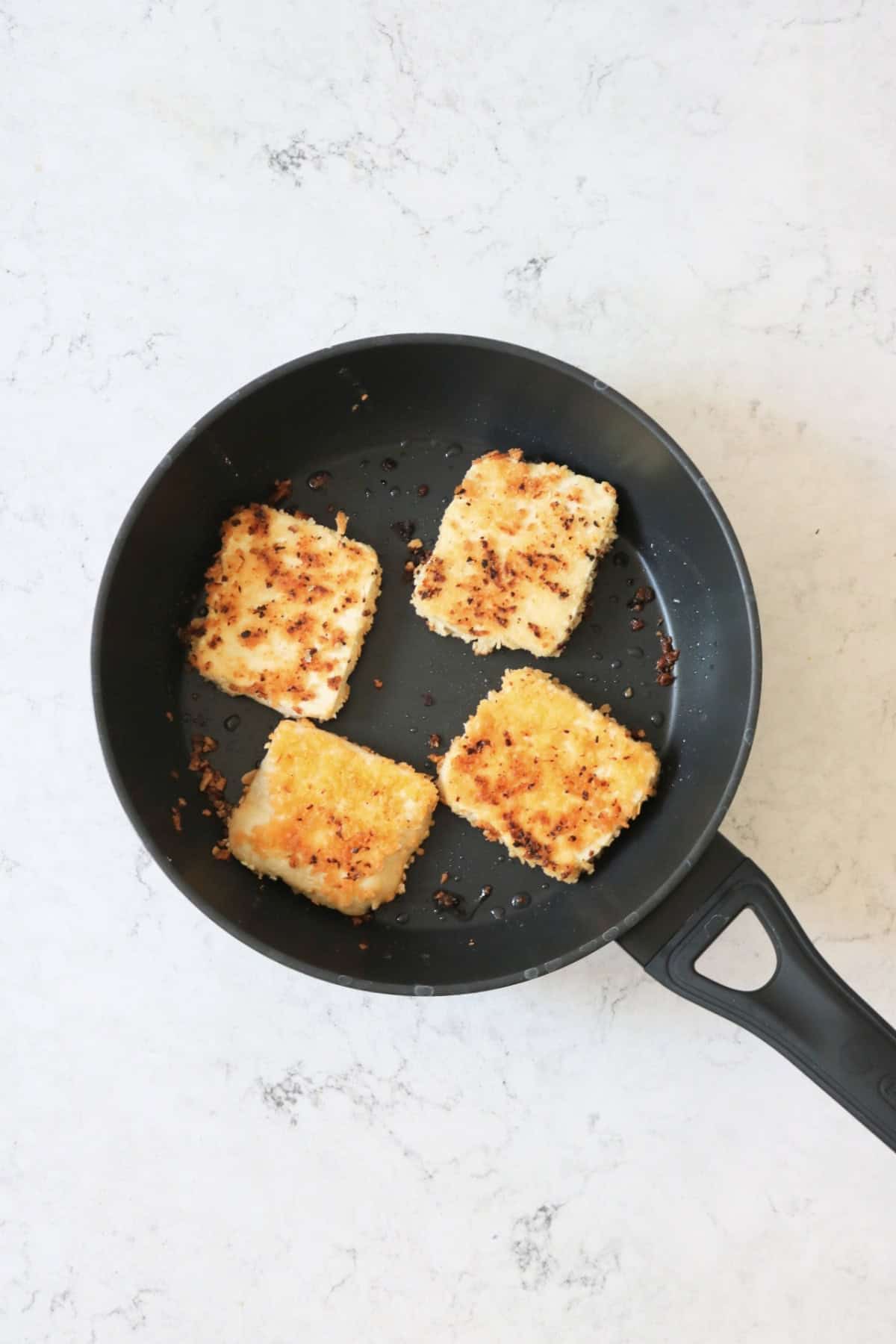 4 slices of tofu coated in lemon-pepper breadcrumbs frying in a large frying pan.
