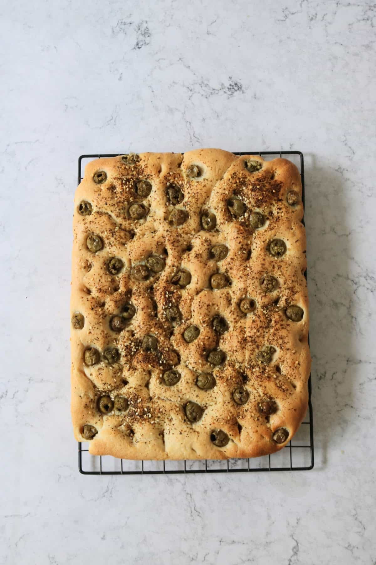 Green Olive and Za'atar Focaccia cooling on a wire rack on top of a white marble counter