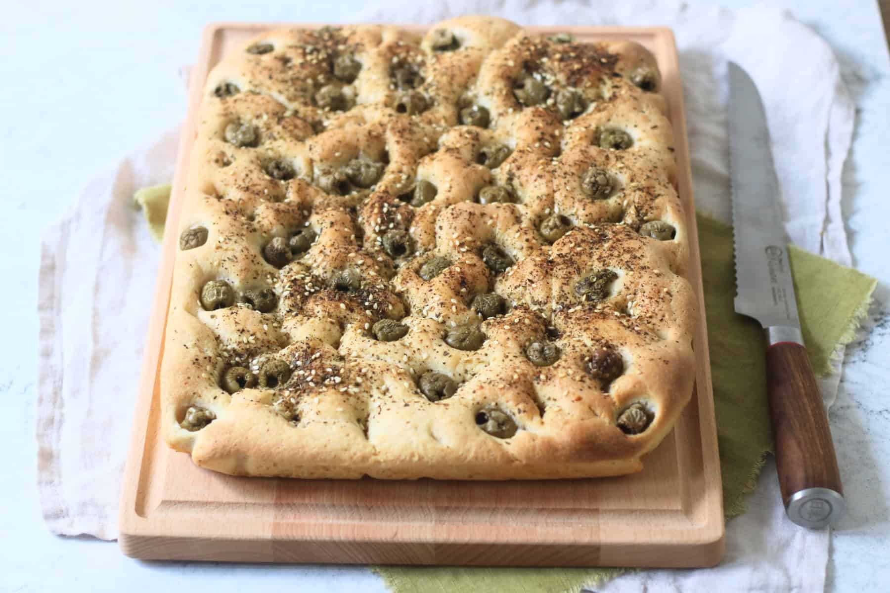 Green Olive and Za'atar Focaccia on a wooden chopping board with a large bread knife