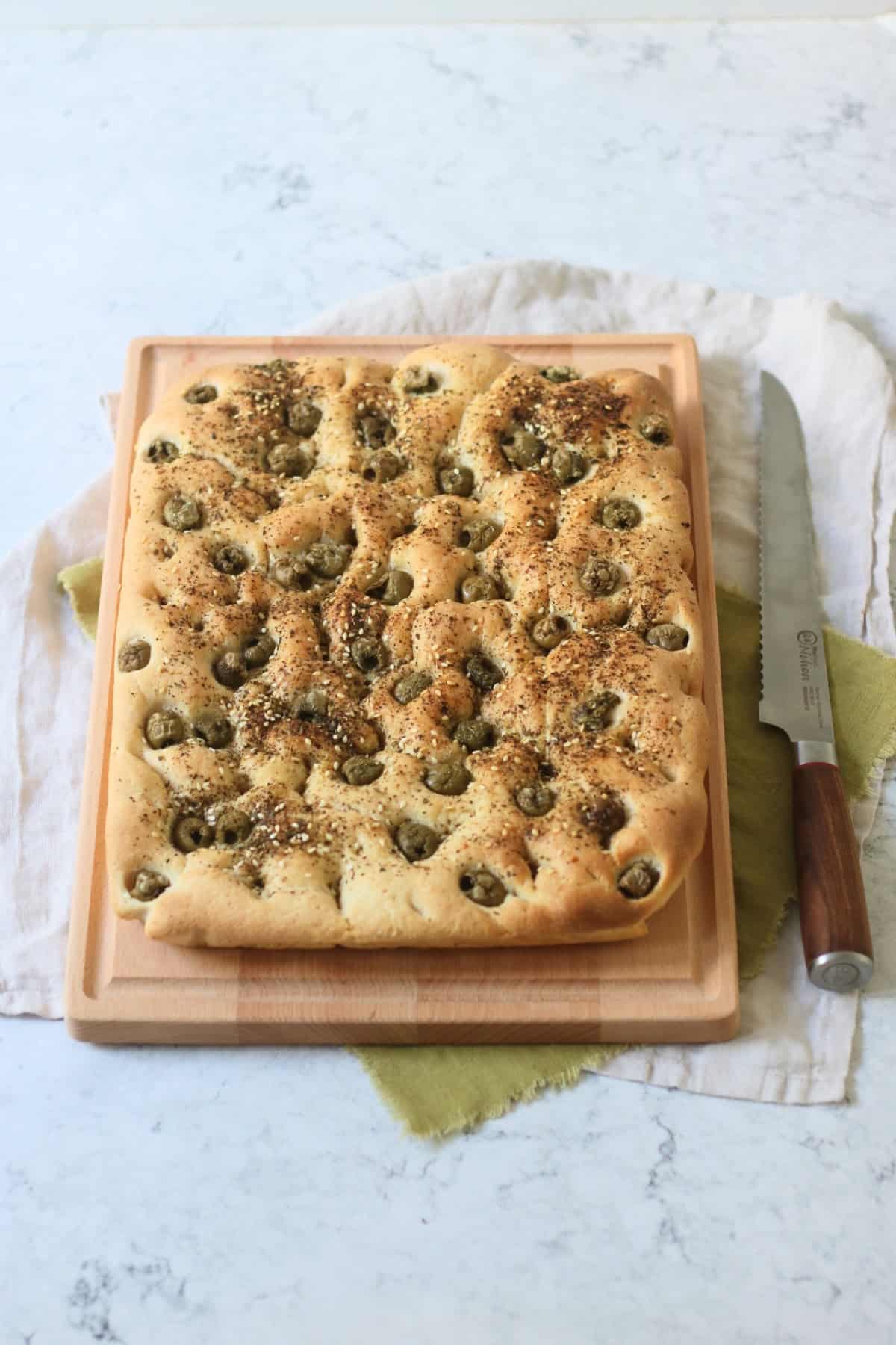 Green Olive and Za'atar Focaccia on a wooden chopping board with a large bread knife