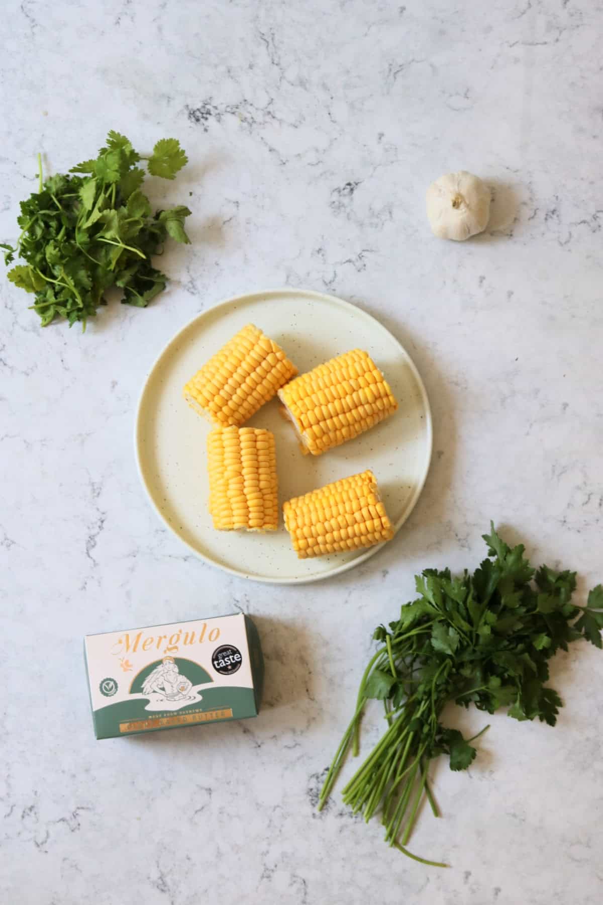 A bunch or coriander, a plate of corn on the cobs, a bulb of garlic, plant based butter and a bunch of parsley, all laid out on a white marble counter top.