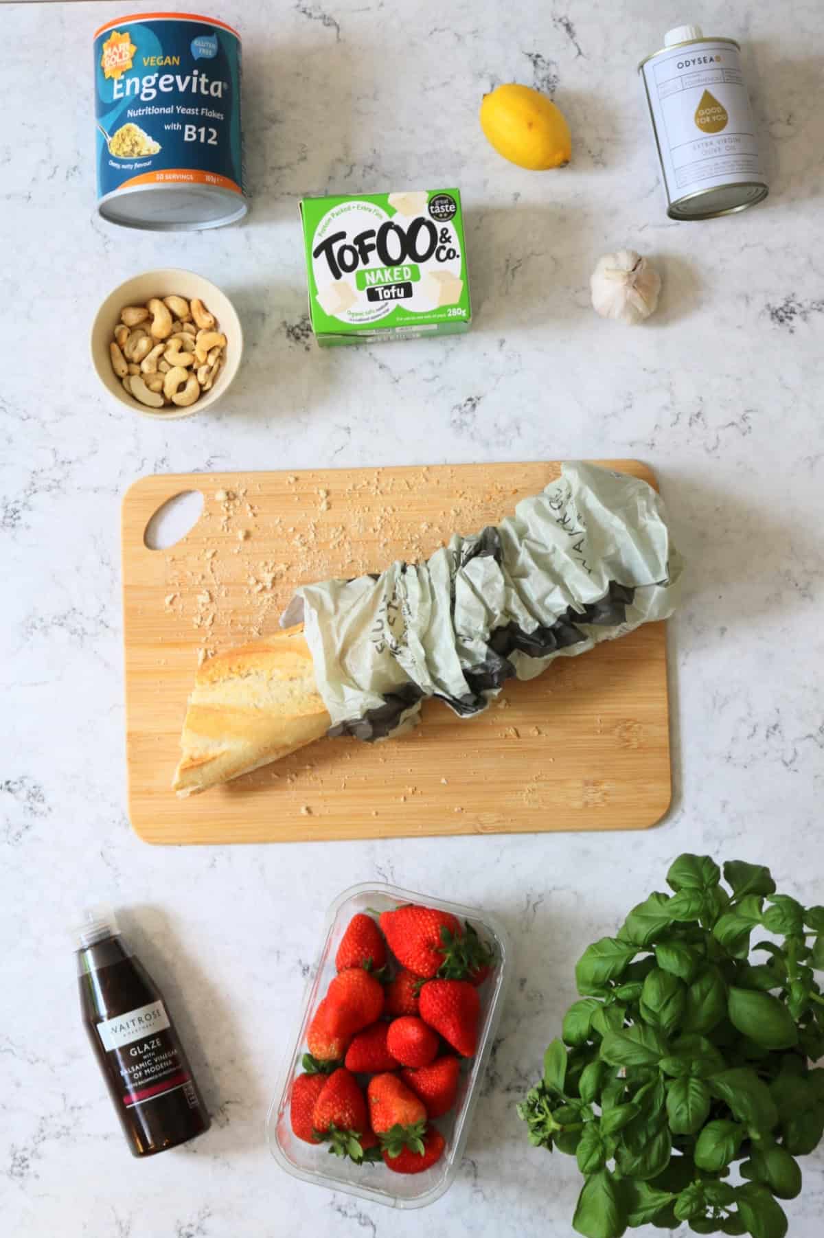 Nutritional yeast, a lemon, olive oil, cashew nuts, tofu, garlic, a baguette, balsamic glaze, strawberries and basil all laid out on a white marble counter top.
