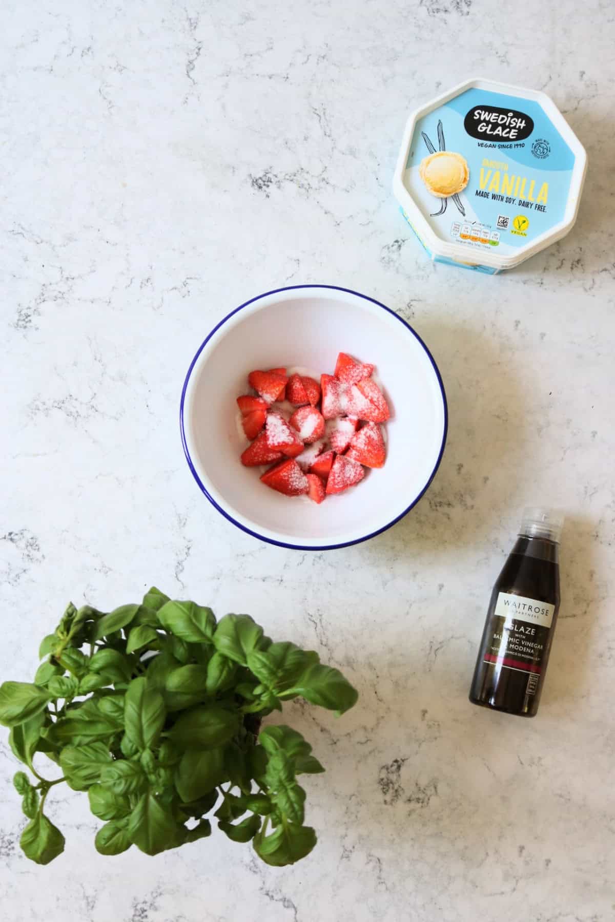 A tub of dairy free vanilla ice cream, strawberries in a bowl with caster sugar, a basil plant and a bottle of balsamic glaze all laid out on a white marble counter top