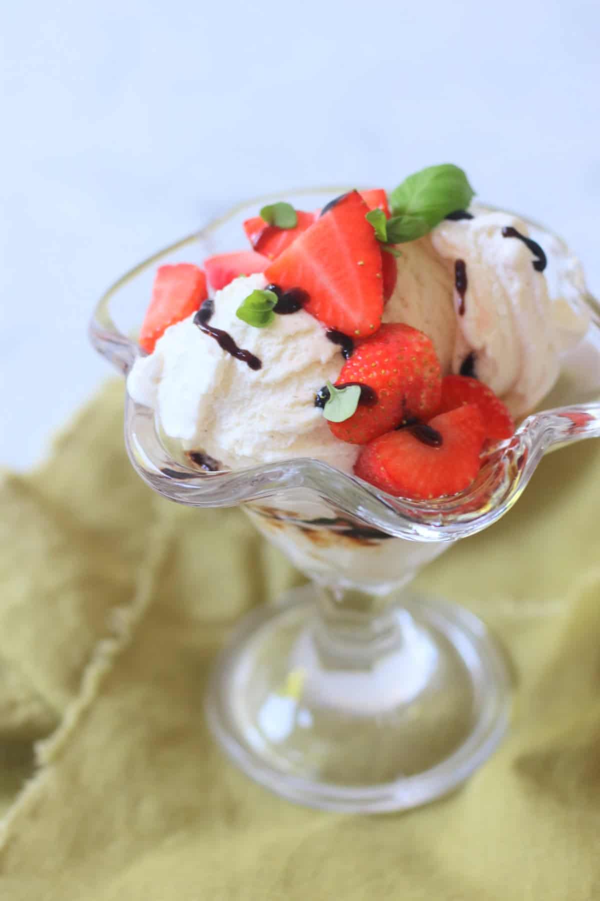 A Vegan Strawberry and Balsamic Ice Cream Sundae made up of layers of balsamic glaze, dairy free ice cream, macerated strawberries and basil leaves served in an ice cream sundae glass on top of a green napkin.