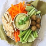 A crudites platter made up of pitta, carrots, celery, falafel, cucumber and orange pepper with a bowl of Roasted Garlic and Basil Hummus