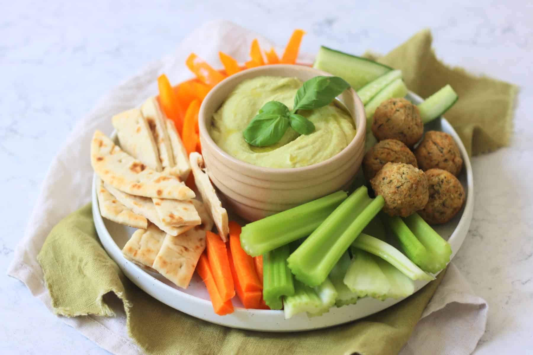 A crudites platter made up of pitta, carrots, celery, falafel, cucumber and orange pepper with a bowl of Roasted Garlic and Basil Hummus